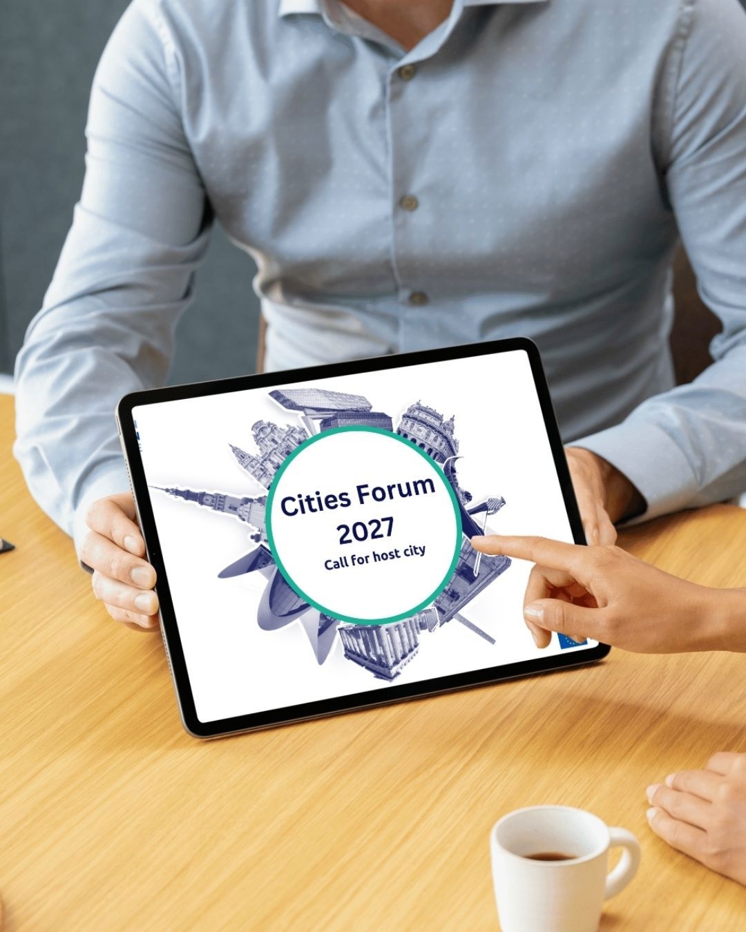 Pessoa a segurar um tablet com o destaque “Cities Forum 2027 – Call for host city”, ilustrado com marcos urbanos europeus, simbolizando a abertura de candidaturas para a cidade anfitriã do Cities Forum 2027.